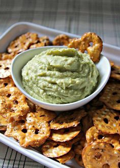 a bowl of guacamole surrounded by crackers on a tray with other snacks