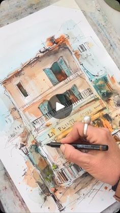 a person is holding a pencil and drawing a house with watercolors on paper