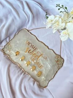 a white and gold tray that says home sweet home next to some flowers on a bed