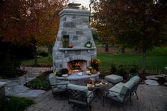 an outdoor fireplace surrounded by chairs and tables in the evening sun with fall foliage around it