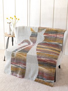 a crocheted blanket on a white chair with flowers in the corner and a vase behind it