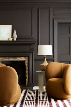 a living room with two chairs and a table in front of a fire place that has a lamp on it