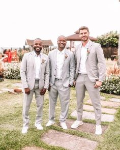 three men standing next to each other in front of a house with flowers on the ground