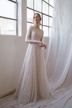 a woman standing in front of a window wearing a long sleeved dress with sequins