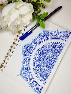 a blue and white drawing on top of a spiral notebook next to flowers with two pens