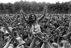 a woman standing in front of a crowd holding her hands up to the sky and smiling