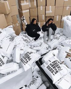 two women sitting on cardboard boxes surrounded by white bags and packages with black lettering that spell out words