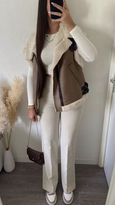 a woman taking a selfie while wearing white pants and a brown vest with fur