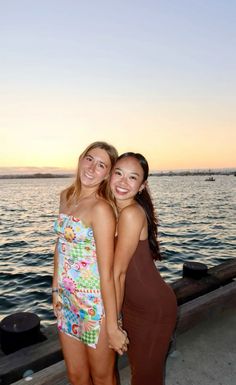 two young women standing next to each other near the water at sunset or sunrise, posing for a photo