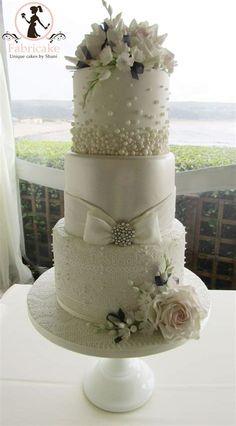 a three tiered wedding cake with white flowers and pearls on the top is displayed in front of a window