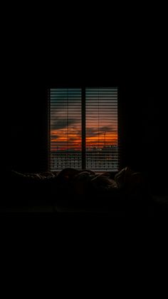 the sun is setting in front of a window that has blinds on it and there is a bed with pillows