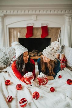 two women sitting on a bed with christmas decorations and cups in front of the fireplace