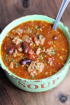 a green bowl filled with chili and beans on top of a wooden table next to a spoon