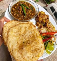 a person holding a plate with some food on it and a bowl of soup in the background