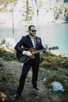 a man in a suit and sunglasses playing an acoustic guitar on the side of a mountain