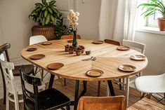 a wooden table with white chairs and plates on it in front of a potted plant