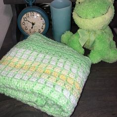 a green stuffed animal next to a crocheted blanket and clock on a table