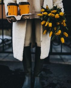 a person in white coat holding two cups and yellow flowers on the other side of their jacket