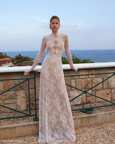 a woman in a long white dress standing on a balcony next to the ocean and looking at the camera