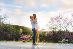 a man and woman kissing in the middle of an empty road with pink flowers behind them