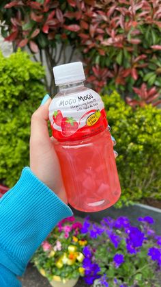 a person holding up a bottle of juice in front of some plants and flowers on the ground