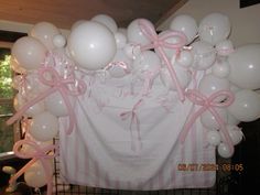 a bunch of white balloons with pink bows and ribbons hanging from the ceiling in front of a wall