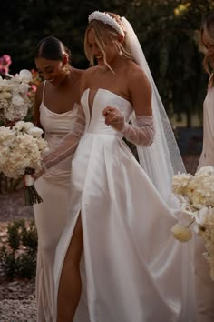 two women in wedding gowns standing next to each other and one is holding flowers