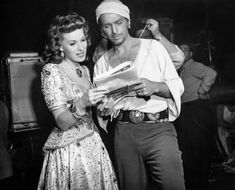 a man and woman standing next to each other in front of a tv screen, reading a book