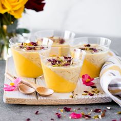 four glasses filled with dessert sitting on top of a wooden tray next to flowers and spoons