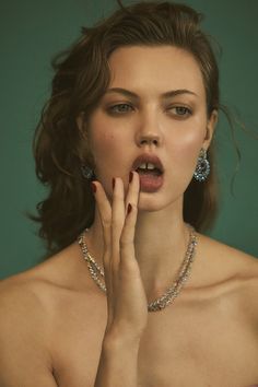 a woman with her hands to her mouth and wearing jewelry on her chest, standing in front of a green background