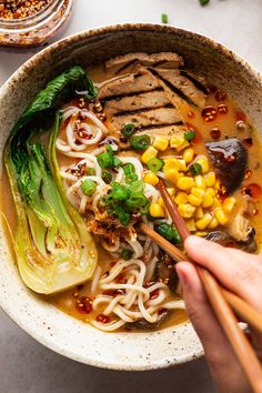 someone is holding chopsticks over a bowl of noodles and meat with corn on the side