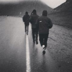 black and white photograph of three people running down the road in the rain with an umbrella over their head