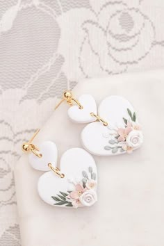 two white heart shaped earrings with pink flowers on them sitting on a piece of fabric