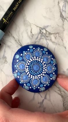 a hand holding a small blue and white painted rock next to a black marker on a marble surface