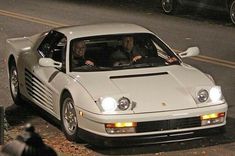 two people in a white sports car driving down the street at night with one person sitting in the driver's seat