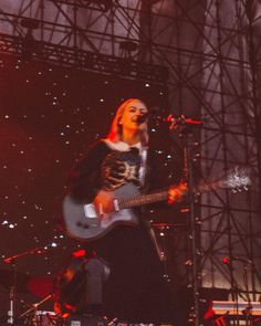 a woman is playing guitar on stage with other people in the background and lights behind her