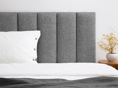 a bed with a gray headboard next to a night stand and flowers in a white vase