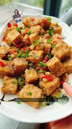 someone is holding a white plate with tofu and vegetables on it in front of a window