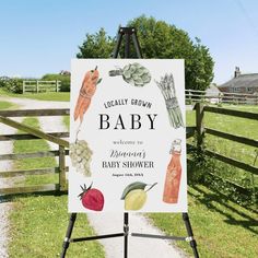 a baby shower sign in front of a wooden fence with vegetables and fruits on it