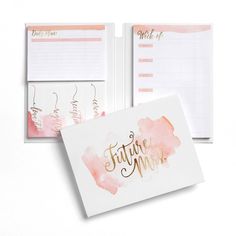 a pink and white planner with gold foil lettering on the cover, next to a notepad