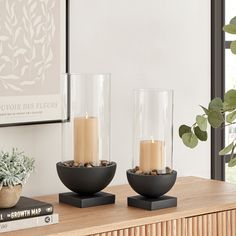 two glass vases with candles on top of a wooden table next to a plant