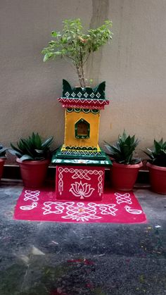 a yellow and green birdhouse sitting on top of a red rug next to potted plants