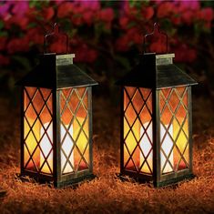 two lit lanterns sitting in the grass with flowers behind them on a sunny day, one is yellow and the other is red