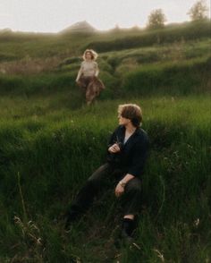 two people are sitting in the grass with one person on his back and another standing behind them