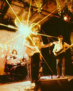 two men are playing guitars on stage with other people in the background and one man is singing