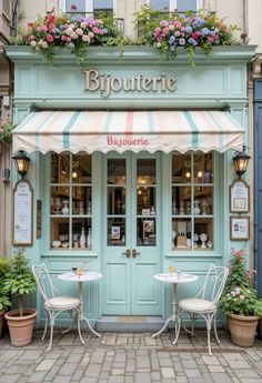 the outside of a restaurant with two tables and chairs in front of it, surrounded by potted plants