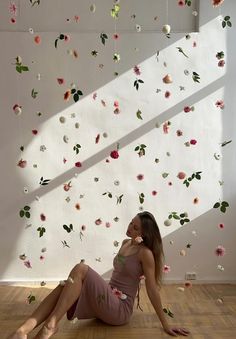 a woman sitting on the floor in front of a wall with flowers hanging from it