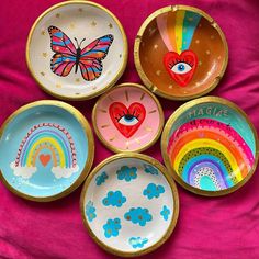 six colorful bowls with designs on them sitting on a pink cloth covered tablecloth, one has a butterfly and the other has a rainbow