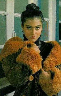 a woman wearing a brown coat and leopard print scarf standing in front of a window