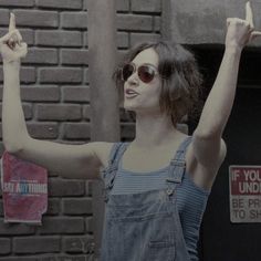 a woman in overalls and sunglasses holding up two fingers while standing next to a brick wall